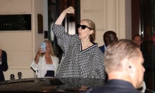 Celine Dion stands next to a car, holding her fist up in the air and cheering.