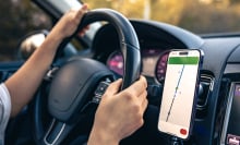 A person's hands on a car steering wheel. A phone with GPS navigation on the screen is attached to the dashboard.