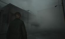 A person with short hair, wearing a dark coat, is standing on a foggy, deserted street. The atmosphere is eerie, with thick mist obscuring much of the surroundings, including power lines and dimly visible buildings in the background. The scene evokes a sense of isolation and tension