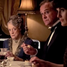 Three characters from 'Downton Abbey' sit at a dinner table. Maggie Smith's character, Dowager Countess, looks at the camera.