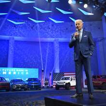 Old white man in blue suit speaks into a microphone. In the background is a display lined with electric vehicles in red, white, and blue variations