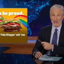 A man sits behind a talk show desk. In the top-left is a Burger King advertisement for Pride.