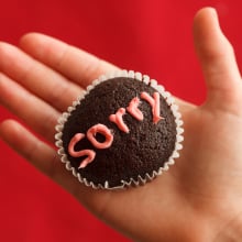 A muffin in the palm of a person's hand with the word "sorry" on it written in frosting