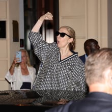 Celine Dion stands next to a car, holding her fist up in the air and cheering.