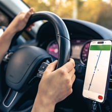 A person's hands on a car steering wheel. A phone with GPS navigation on the screen is attached to the dashboard.