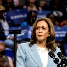 Vice President Kamala Harris looking serious in a pale blue suit at a campaign rally.