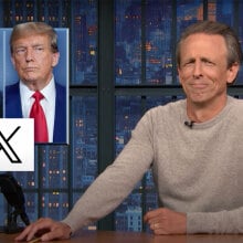 A man sits behind a talk show desk. In the top-left is an image of two other men.