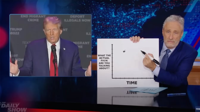 A man sits behind a talk show desk holding a chart. In the top-left is an image of another man wearing a suit.