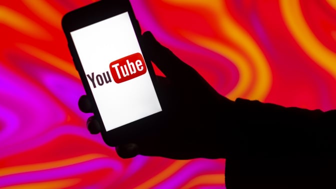 A silhouette of a person holding a phone displaying the YouTube logo surrounded by pink and orange tie dye. 