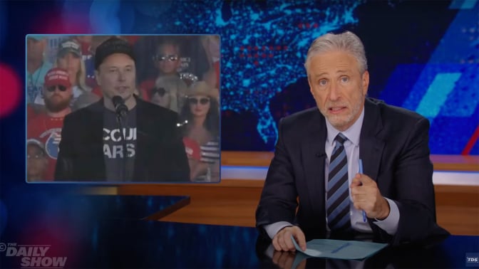 A man in a suit sits behind a talk show desk. In the top left is an image of another man in a dark baseball cap with a crowd behind him.