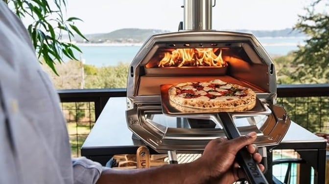 a pizza going into an ooni oven with a fire lit. the background is a beautiful body of water and mountain in the distance.