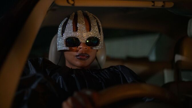 A woman drives a car wearing sunglasses and a sequined hat.