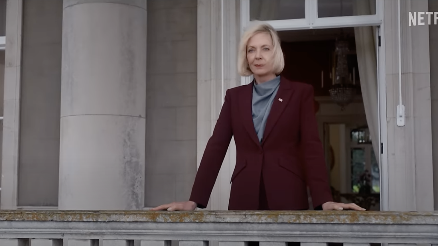 Allison Janney wears a red suit standing on a stone balcony.