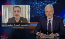 A man in a suit sits behind a talk show desk. In the top-left is an image of a man on the news and the phrase "de-escalation through escalation."