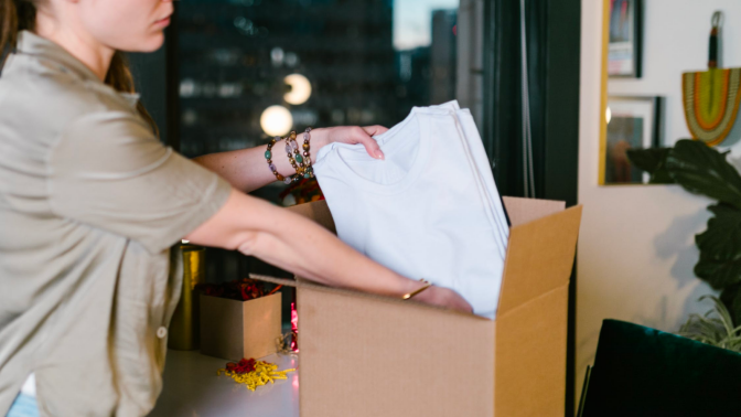 a woman in a tan polo packs a stack of white t-shirts into a cardboard box. there are four beaded bracelets on her wrist